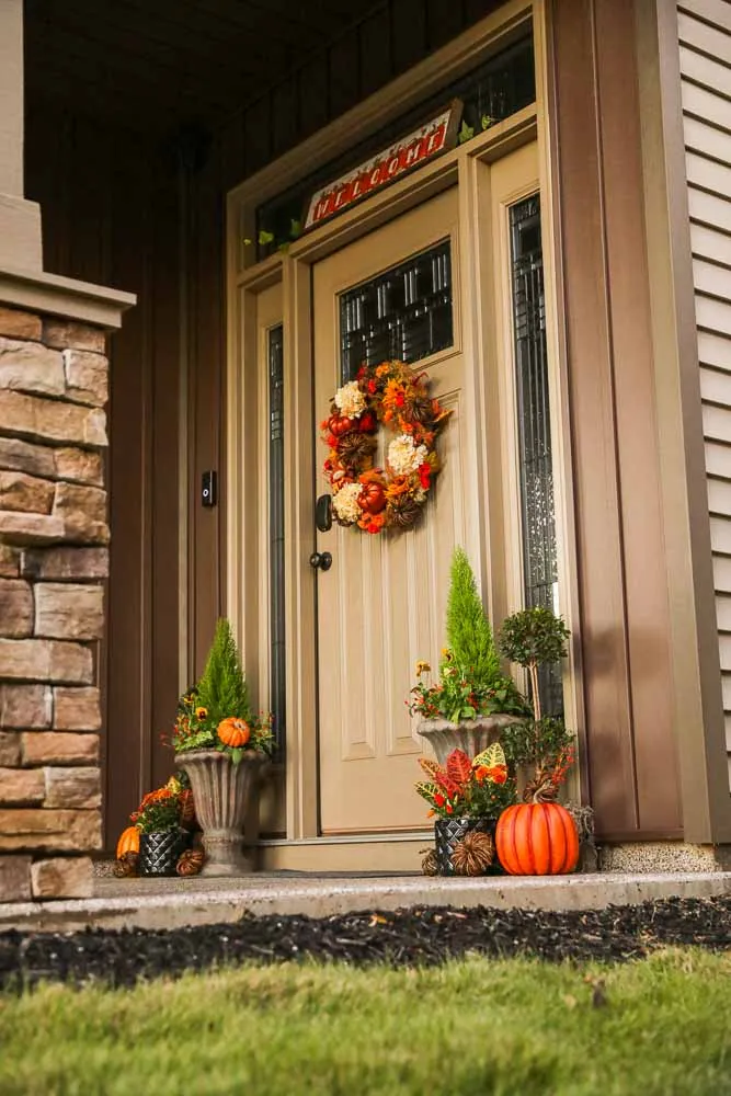 Layered autumn porch entry: cypress topiary and croton in cast urns, ornamental cabbage at the base, pumpkins stepped down to the walk